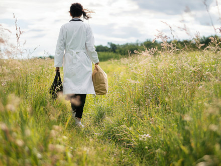 Eine Frau mit Arztkittel trägt zwei Reisetaschen über eine Wildblumenwiese.