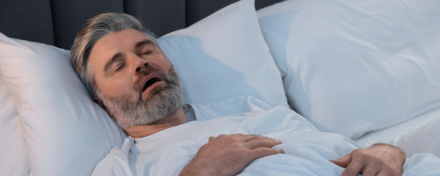 Mittelalter Mann mit grauem Bart schläft in Rückenlage im Bett mit geöffnetem Mund, Hinweise auf mögliche Schlafapnoe oder Schnarchen. Schlafzimmerumgebung mit weißen Kissen und Bettdecke, Fokus auf Atemprobleme im Schlaf.