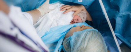 Frau liegt während eines Kaiserschnitts im Operationssaal, hält ihr Neugeborenes zum ersten Hautkontakt im Arm. Das Baby wird von medizinischem Personal unterstützt und liegt geschützt in Tüchern.