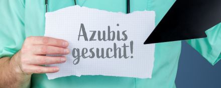 Eine Person im grünen Kasak mit Stethoskop um den Hals hält ein Blatt auf dem "Azubis gesucht" steht. In der anderen Hand hält sie einen schwarzen Pfeil, der auf das Blatt zeigt.