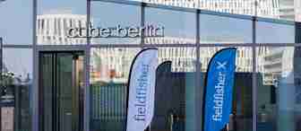 The entrance to Cube Berlin with reflective glass walls, showcasing two blue and white flags bearing the "fieldfisher" logo. The modern building architecture is visible in the background.