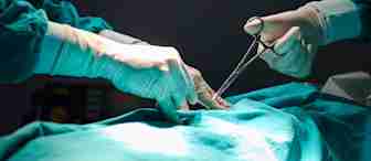 Two surgeons in green surgical gowns and gloves perform a procedure on a patient, using surgical instruments under focused lighting.