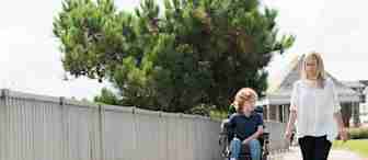 A young boy using a motorized wheelchair and a woman walking beside him are on a paved path next to a fence and trees. The sky is clear and there's a building in the background. Both look towards each other, appearing to be in conversation.