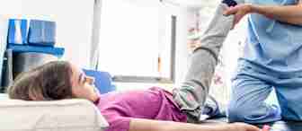 A healthcare professional in blue scrubs holds up a young girl's leg while she lies down on a bed in a brightly lit medical room. The girl, wearing a pink shirt and gray pants, looks up at the professional who appears to be using a device on her leg.