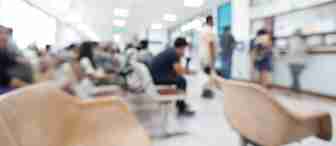 Blurry image of people sitting in a waiting area, possibly in a hospital or clinic, with rows of chairs and a reception counter in the background. The scene looks busy and well-lit.