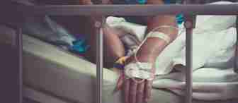 A person lies in a hospital bed with an IV drip attached to their arm. The person looks weary, with one hand resting on the bed railing and the other covering their face. The surroundings are dimly lit, emphasizing a somber mood.