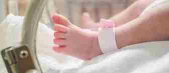 Close-up of a newborn baby's foot with a hospital identification band around the ankle, resting inside an incubator. The scene is softly lit, highlighting the baby's delicate skin and creating a warm and gentle atmosphere.