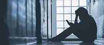 Silhouetted person sitting on the floor in a narrow hallway, leaning against a wall with one arm resting on their knees, looking at a smartphone. Light coming through a window at the end of the hallway creates a moody atmosphere.