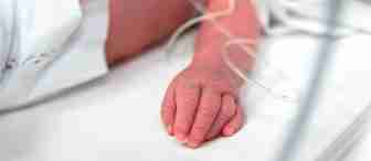 Close-up of a newborn baby's hand and arm, with medical tubes attached, resting on a white hospital bed. The baby is partially covered with a diaper, indicating a clinical or neonatal setting.
