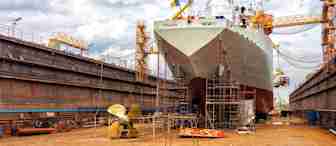 A large ship is docked in a dry dock undergoing maintenance or construction. The ship is surrounded by scaffolding and various equipment. The sky is cloudy and overcast. Yellow cranes are visible on either side of the ship, and the ship's propeller is visible on the ground in front.