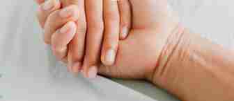 A close-up of two hands clasped together. One hand has a medical IV line attached, suggesting a hospital setting. The other hand is holding it gently, conveying a sense of support and care. Both hands rest on a light-colored bed sheet.