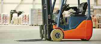 A red and black forklift is parked indoors on a concrete floor. In the background, there are stacks of boxes and pallets against a large industrial wall. The warehouse setting is well-lit.