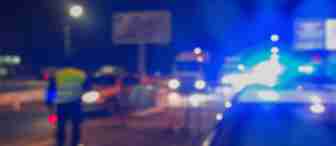 Blurred image of a nighttime scene with several cars on the road, flashing blue and red lights from police vehicles, and a person in a reflective vest standing by the roadside. The atmosphere suggests an ongoing emergency or traffic stop.