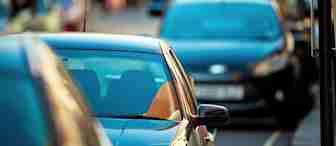 A row of parked cars lines a city street, with the foreground car in sharp focus and the background cars and buildings blurred.