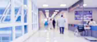 A blurred image of a hospital corridor with bright lighting. People are walking in the distance, and a wheelchair is positioned near a counter on the right side. Large windows on the left let in natural light. Tables with information are on the right side of the hallway.