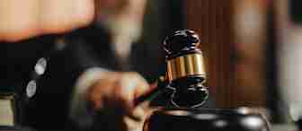 A judge in a courtroom holds a wooden gavel, poised to strike it on the sound block, with the judge’s face blurred in the background.