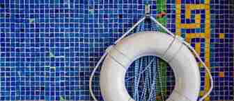 A white lifebuoy with blue ropes is mounted on a wall covered in blue mosaic tiles with yellow, green, and orange geometric patterns on the right side.