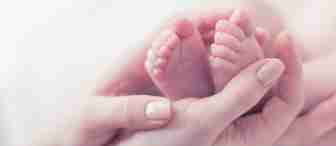 Close-up of an adult's hands gently holding a baby's feet. The image focuses on the tiny toes and the tender, delicate interaction, highlighting the softness and innocence of the baby. The background is blurred and light.