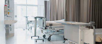 A hospital room with multiple empty beds, each equipped with adjustable bed trays. The room features medical equipment, a sink on the left wall, and light flooding in from a large window in the background. Curtains hang between the beds for privacy.