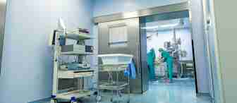A hospital operating room with medical equipment on carts. Two surgeons in green scrubs and surgical caps are performing a procedure near an operating table in the background. The room has sterile blue walls and bright lighting.