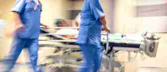 Blurred motion image of medical staff in blue uniforms pushing a patient on a hospital bed down a hallway, indicating urgency. The patient is laying down, and the surroundings are typical of a medical facility, reflecting a fast-paced emergency scenario.