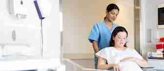 A pregnant woman sits in a wheelchair, holding her belly, while a nurse in blue scrubs stands behind, guiding the wheelchair. They are in a hospital room with medical equipment and an empty bassinet nearby.