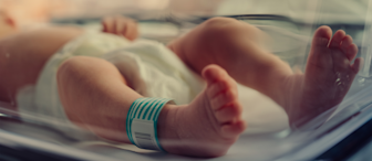 A newborn baby lies in an incubator, wearing a diaper and an identification wristband. The baby's feet are in focus, slightly elevated, and toes are clearly visible. The background is soft and out of focus, emphasizing the fragile and delicate state of the infant.