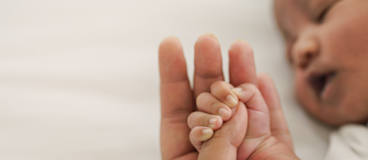 Close-up of a baby holding an adult's finger while resting on a white surface. The baby's small hand is grasping the adult's finger, showcasing the contrast in size and the bond between them. The baby's face is partially visible in the background, appearing to rest peacefully.
