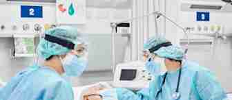 Two healthcare workers in full protective gear, including face shields, masks, gloves, and gowns, attend to an elderly patient lying in a hospital bed. Medical equipment and monitors are visible in the background. The environment appears to be an ICU.