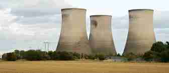 Three large, cylindrical cooling towers stand against a cloudy sky. They are surrounded by greenery and a field in the foreground. The towers are part of an industrial plant, possibly a power station.