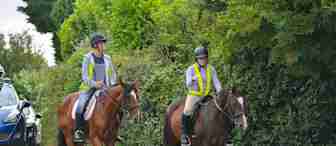 Two people, wearing helmets and reflective vests, are riding brown horses along a quiet, paved road bordered by lush green hedges. In the background, a parked blue car is partially visible. The riders appear to be relaxed and enjoying their leisurely ride.