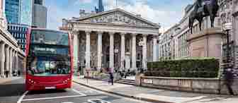 A red double-decker bus with the destination "London Bridge" displayed on its sign passes in front of the Royal Exchange in London. Modern office buildings and a statue of a man on horseback are visible in the background under a partly cloudy sky.