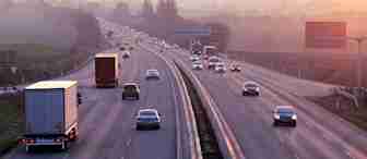 A wide highway with multiple lanes in each direction is pictured during sunset. Several cars and trucks are driving on both sides of the road, with a scenic countryside and trees in the background. The sky displays beautiful hues of pink, purple, and orange.