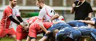A group of rugby players in red and white jerseys scrum against another group in blue jerseys on a grassy field. A person in black clothing stands nearby, holding a whistle. The players are intensely engaged in the match.