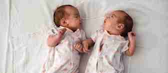 Two newborn babies lie on their backs on a white sheet. They are dressed in matching white onesies with pink polka dots and small fox illustrations. The babies face each other, with their heads slightly turned towards one another. One baby has a hand raised.