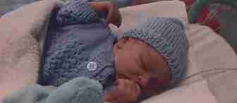 A newborn baby rests in an incubator, wearing a blue knitted hat and matching sweater. The baby has a nasal tube for oxygen and is covered with a white blanket. A soft blue toy is near its small hand. The setting appears to be a neonatal care unit.