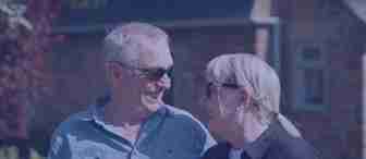 An elderly couple wearing sunglasses shares a warm smile while standing outside on a sunny day. The man is wearing a light blue shirt, and the woman is dressed in black with short blonde hair. They are in front of a brick building with greenery in the background.