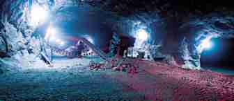A large underground cave is illuminated by bright lights, revealing construction machinery, equipment, and piles of rocks. Tire tracks are visible on the ground, indicating heavy vehicle activity. The cave has a rugged, rocky ceiling and walls.