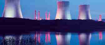A nighttime scene of a nuclear power plant with four illuminated cooling towers and reflected in a calm body of water in the foreground. The towers emit a soft pink and blue glow against the evening sky, creating a serene and surreal atmosphere.
