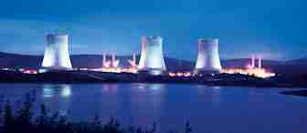 A nuclear power plant with four cooling towers illuminated at dusk. The plant is situated near a body of water with lush greenery in the foreground and mountains in the background, all under a twilight sky.