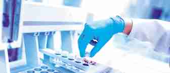 A scientist wearing blue gloves is placing a small vial into a machine filled with multiple vials in a lab. The setting is brightly lit and appears sterile, emphasizing the scientific nature of the work being conducted.