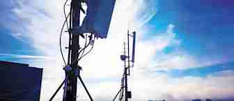 Silhouettes of telecommunications towers with various antennas and equipment are seen against a bright, cloudy sky at sunset or sunrise. The scene is set atop a building in an urban area.