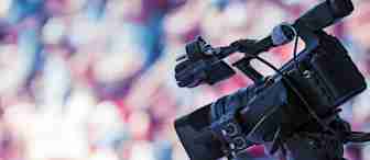 A professional video camera on a tripod is focused on an event. The background is blurred with colorful, out-of-focus shapes, suggesting a vibrant setting with a crowd or audience. The camera is equipped with various attachments and appears ready for filming.