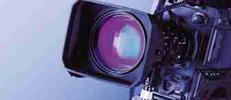 Close-up of a professional video camera lens with various buttons and components visible. The lens has a large, reflective surface, and the camera is positioned against a gradient blue background.