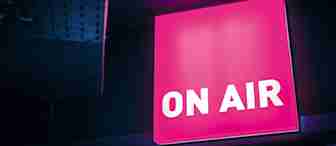 A brightly illuminated pink "ON AIR" sign is mounted on a dark wall, signaling that a live broadcast is in progress. The surrounding area is dimly lit, highlighting the glow from the sign.