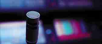 Close-up of a microphone in a dimly lit environment with blurred, colorful screens in the background. The image suggests a high-tech, dynamic setting, possibly related to audio production, broadcasting, or a live performance.