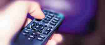 Close-up of a hand holding a black remote control pointed towards a television screen in the background. The TV screen is blurred with a purple hue, and the remote control buttons are slightly out of focus.