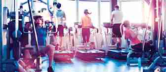 A group of people exercising in a gym. Two men and two women are using treadmills in the background. In the foreground, one man is using a weight machine, and one woman is performing pull-ups. The gym is brightly lit with large windows.
