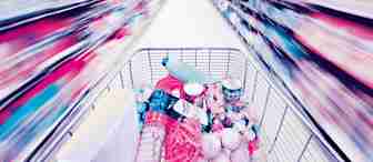 A shopping cart filled with various items, such as bottled water, soda, canned goods, and produce, is being pushed down a supermarket aisle. The image is blurred, giving a sense of fast movement through the brightly lit store.