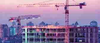 Construction site featuring two large cranes and an unfinished multi-story building under a colorful dusk sky. The background displays a cityscape with various buildings illuminated by the transitioning evening light, creating a blend of urban progress and twilight hues.